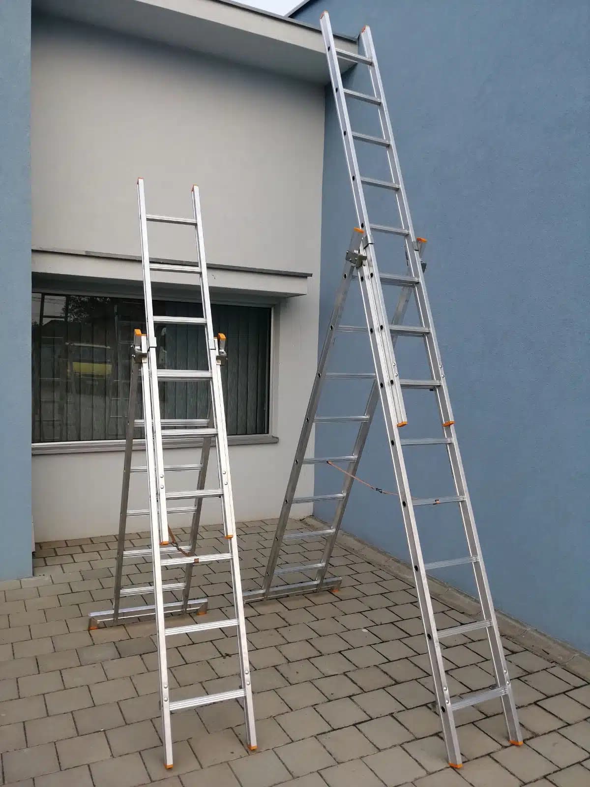 Dvoje aluminijumskih trodelnih merdevina naslonjenih na zid zgrade